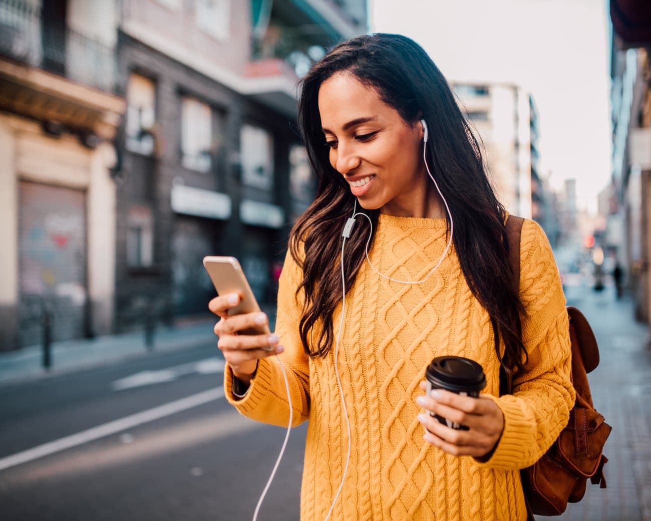 A woman walking down the street with a coffee, checking her phone on the way to a local spot