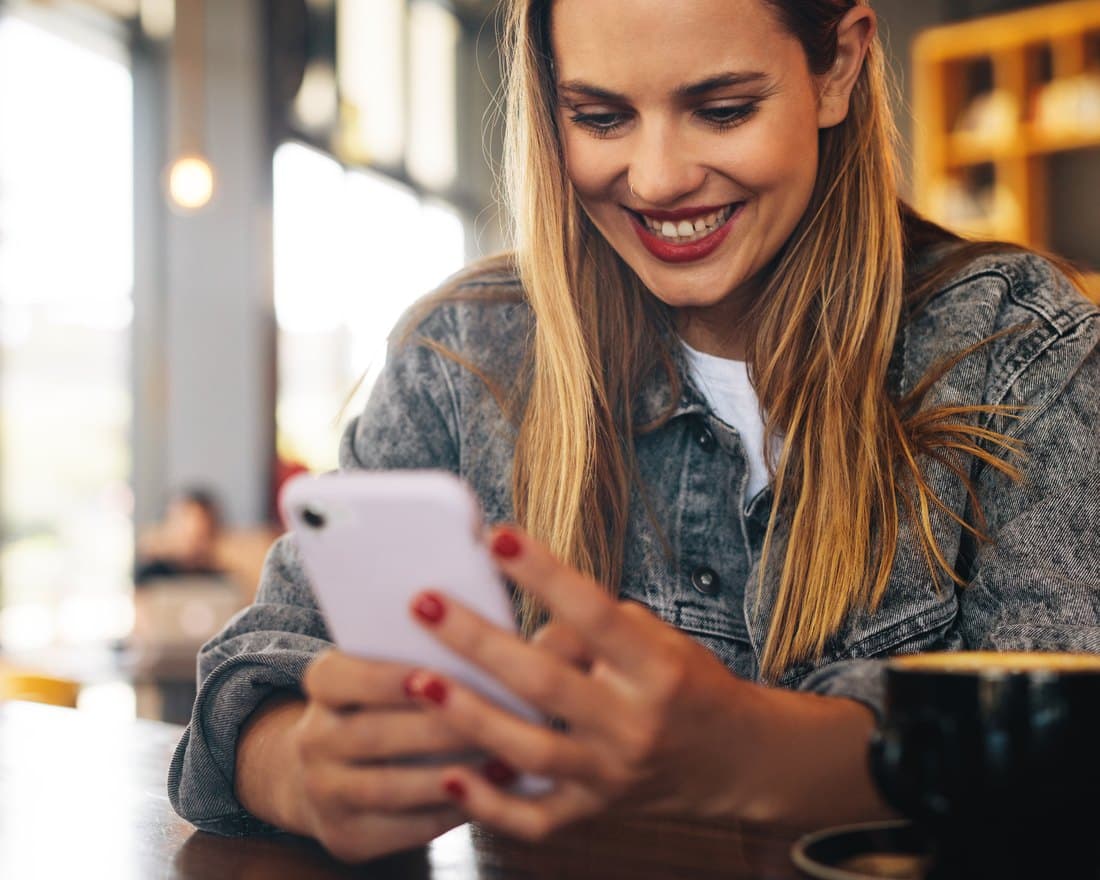 A woman smiling at her phone in a sunlit café after redeeming a reward