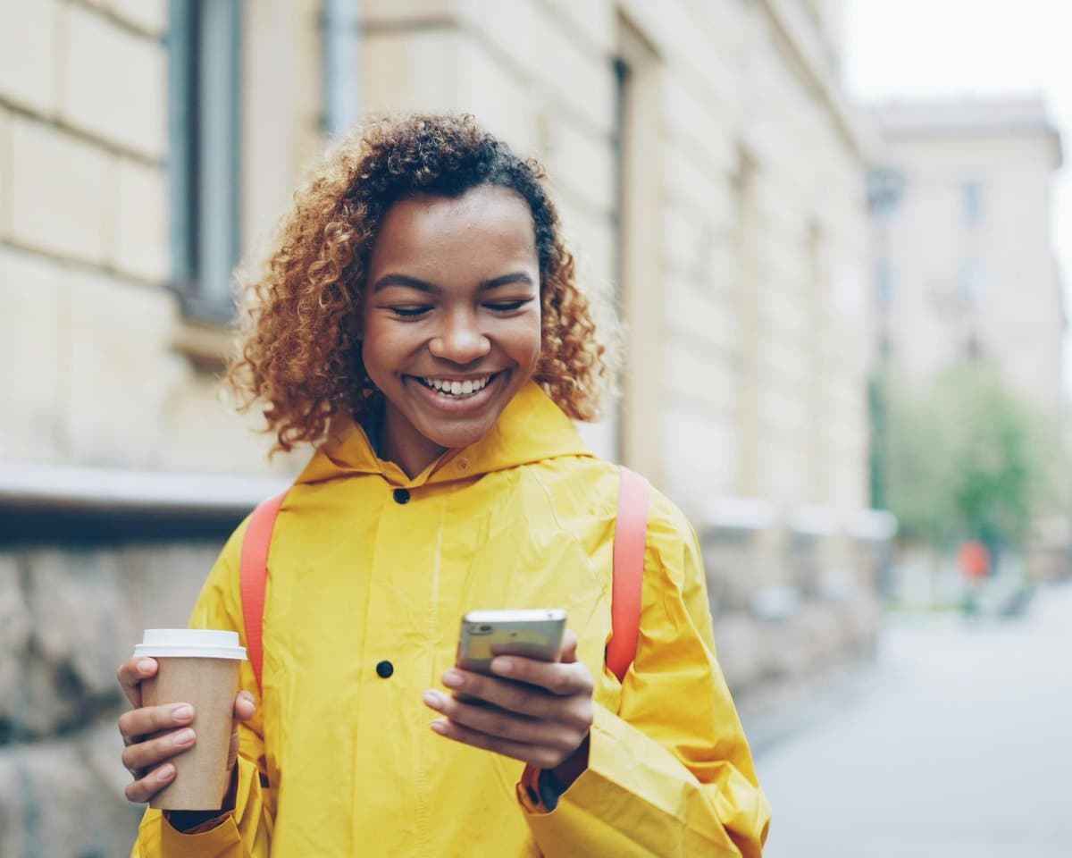 A smiling woman with a coffee checking her phone on a city street